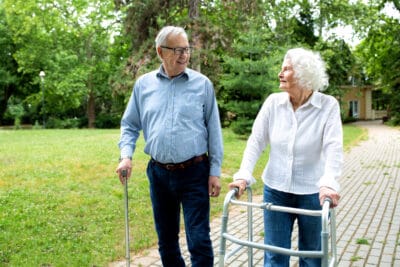 An older man with a cane and an older women with a walker taking a walk outside on a path near green trees.