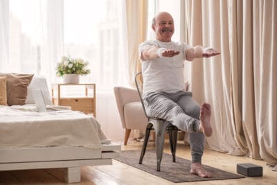 An older man practicing a yoga core exercise seated in a chair in a bedroom with one knee extended and both arms raised in front of hid body.