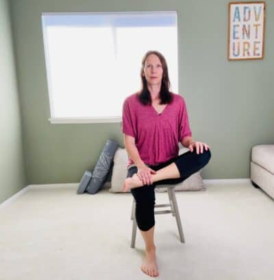 A woman seated in a chair for chair yoga easy pose with one ankle crossed over the opposite knee.