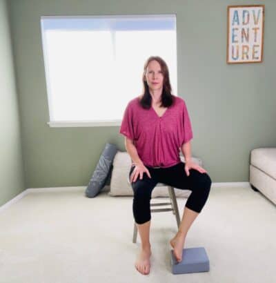 A woman seated in a chair for modified easy pose with one ankle resting on a yoga block for modified easy pose.