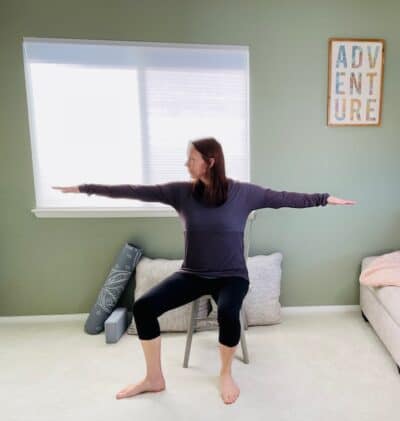 A woman seated in a chair practicing the chair yoga move Warrior 2 with one leg rotated outward and both arms raised to shoulder height.