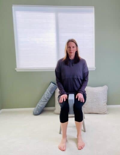 A woman seated in a chair near a window with a yoga block between her knees for a seated hip isometric adduction exercise.