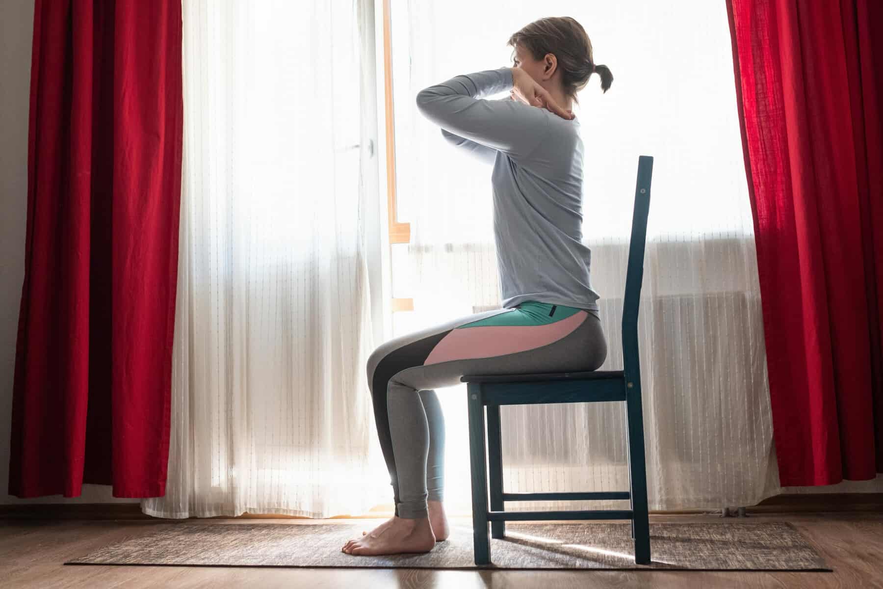 A women in a chair doing an upper body shoulder stretch near a window.