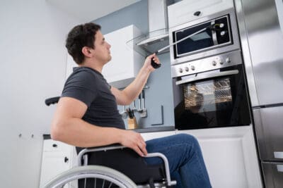 A young man seated in a wheelchair using a long handled reacher to push the buttons on a microwave.