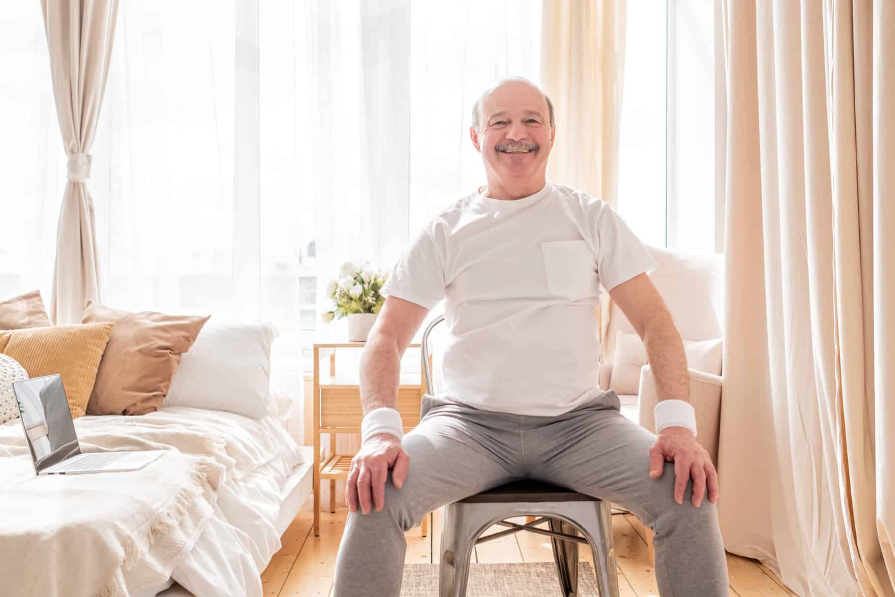 An older man seated in a chair by a sunlight window preparing to practice chair based exercises..