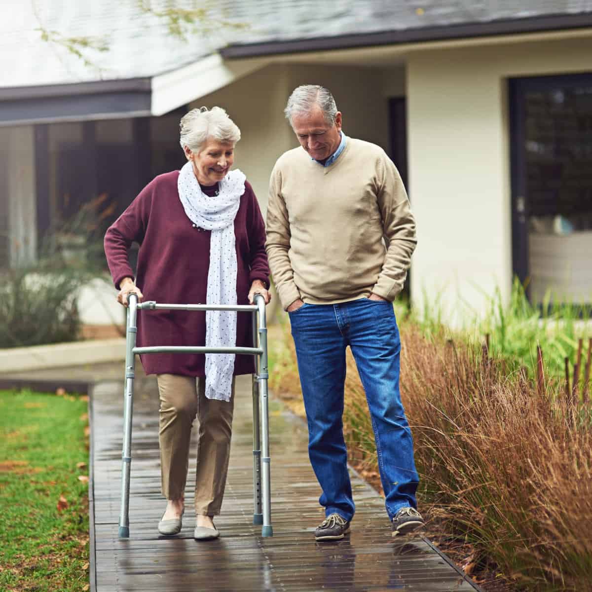 An older woman with a walker walking next to an older man outside a house on a walkway.