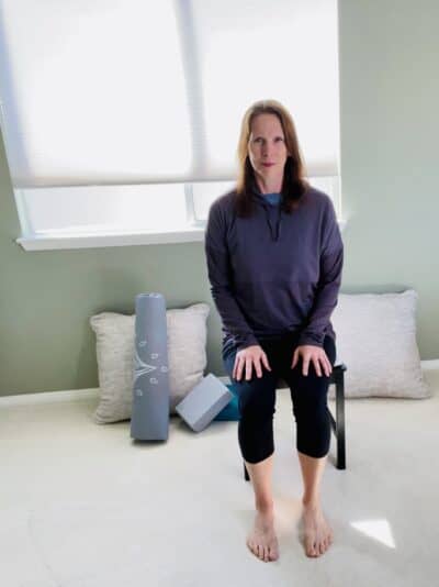 A women seated in a chair near a window with upright posture in preparation for chair yoga exercise.