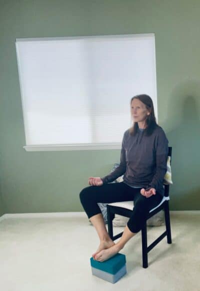 A women seated in a chair performing a restorative yoga pose with feet supported on yoga blocks and the soles of the feet together.