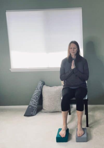 A women seated in a chair performing a restorative yoga pose with her palms together at the heart.