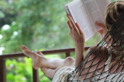 A cropped image of a woman's legs and arms sitting outside in a hammock reading a book