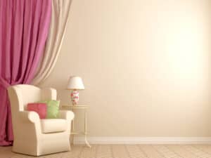 A beige arm chair in a room with beige walls and a pink curtain next to a stand with a lamp