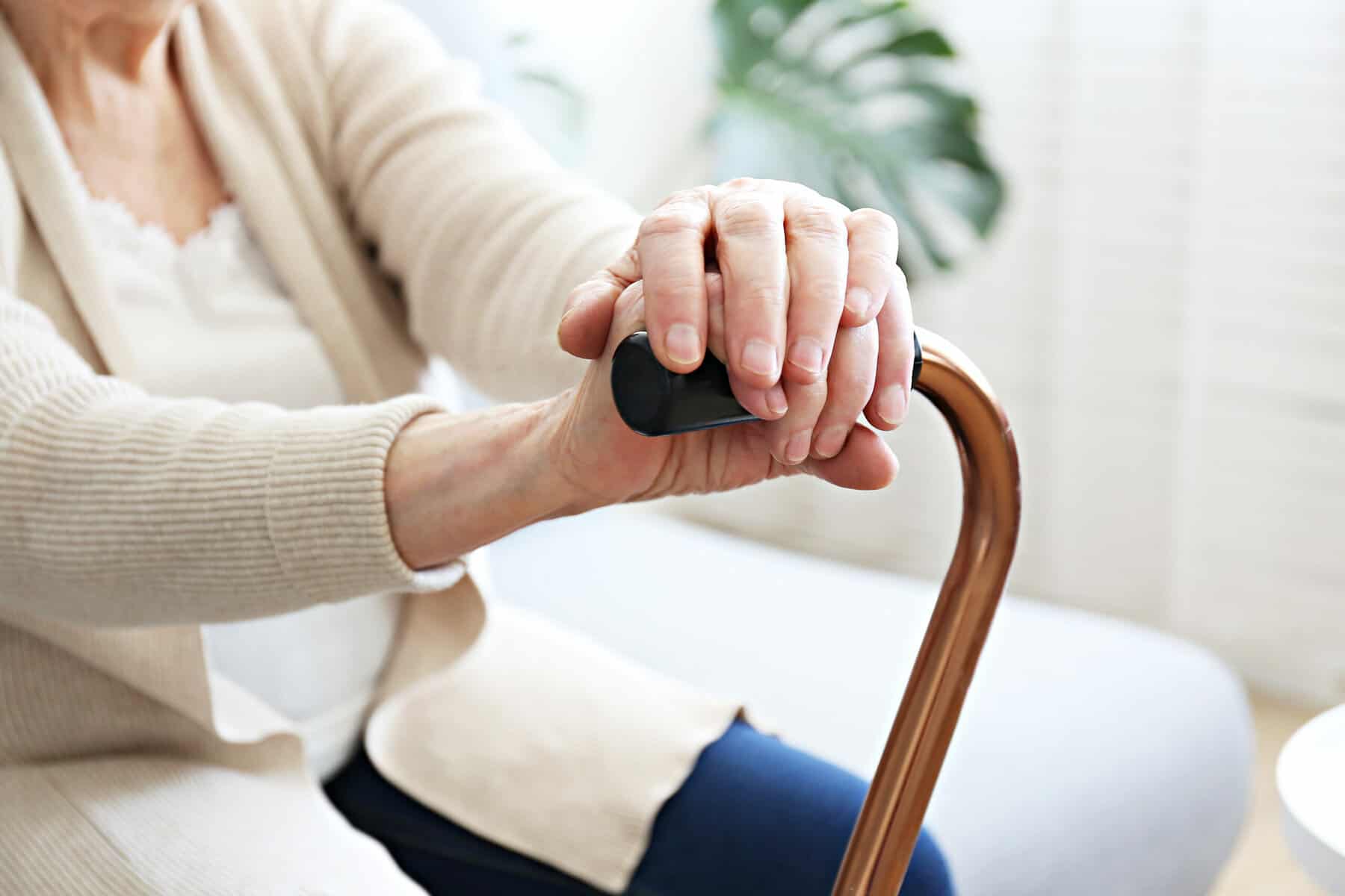 A cropped photo of an older woman seated on a chair holding a cane in her hands.