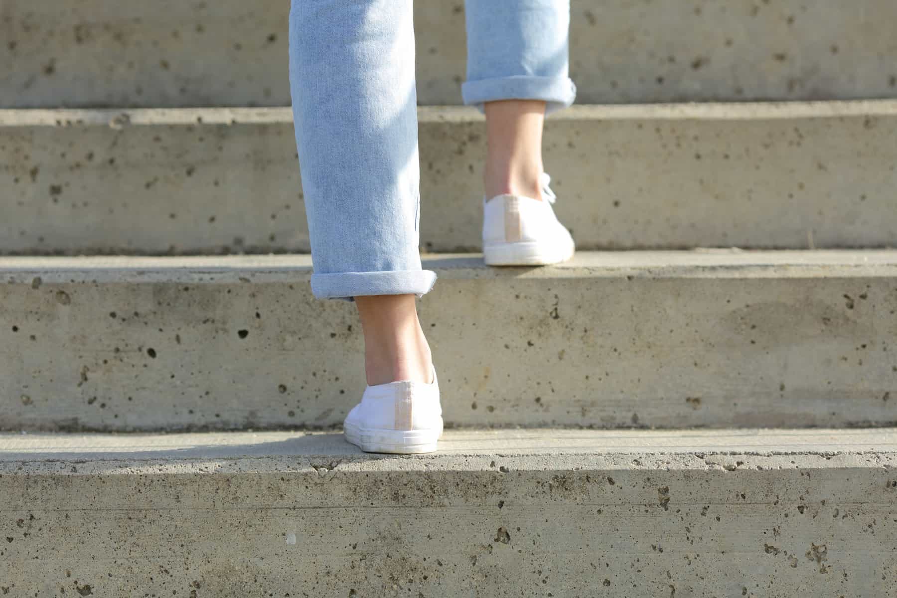 A cropped image of a woman wearing white sneakers and climbing stairs outside.