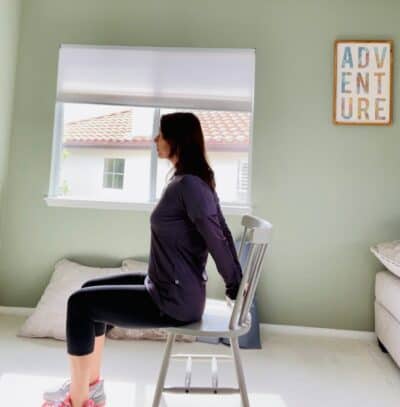 A woman sitting in a chair practicing chair yoga pose bound hand stretch with her hands clasped behind her back.