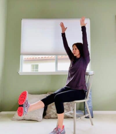 A woman seated in a chair for chair yoga with one knee extended and both arms raised overhead.