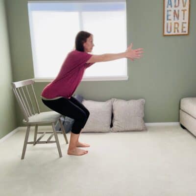 A woman performing a chair yoga chair pose with arms reaching forward and a chair behind her.