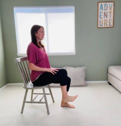 A woman seated in a chair for chair yoga low lunge with one knee bent and raised in the air.