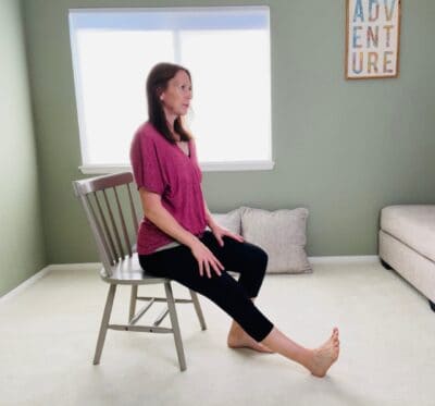 A woman seated in a chair for chair yoga half staff pose with one knee extended and the heel placed on the floor.