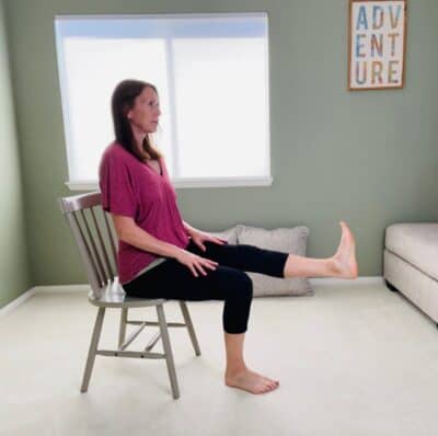 A woman seated in a chair with one knee extended and lifted for seated straight leg raise.