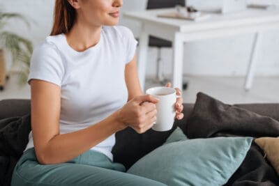 A cropped image of a young woman relaxing while seated on a sofa and holding a white coffee mug.