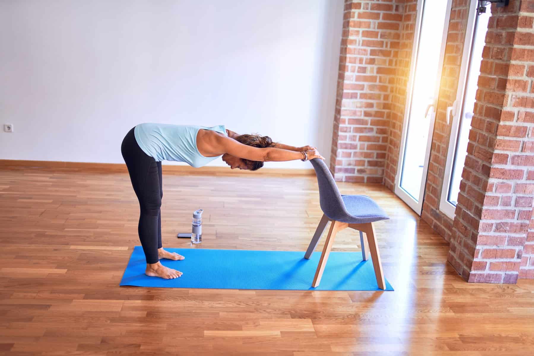 A woman with a light blue shirt and black yoga pants practicing a standing chair yoga position with a chair for support, pose Downward Facing Dog in a sunlit studio with hardwood floors and white and brick walls.