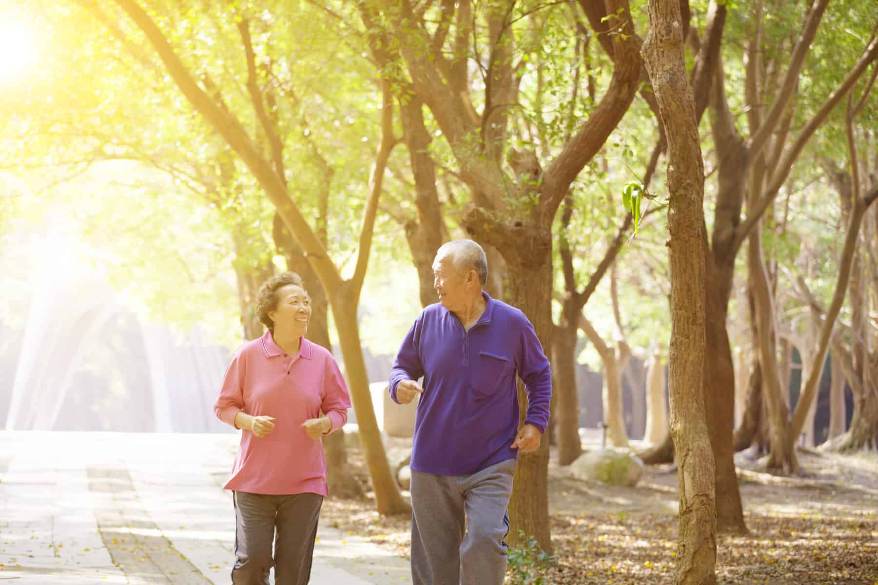 Older adults exercising and walking in the park on a sunny day with trees in the background.