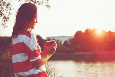 A young woman holding a red coffee mug and relaxing while looking upon a lake at sunrise