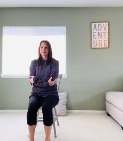 A woman seated in a chair for chair yoga with arms crossed and grasping opposite hand to opposite elbow.