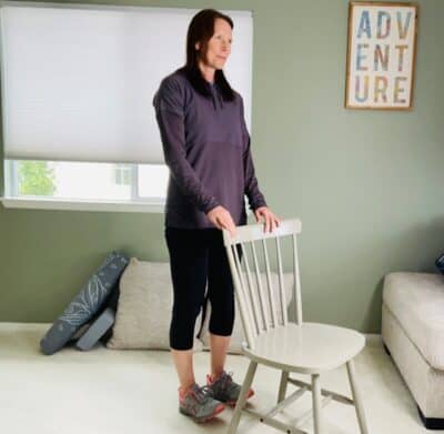 A woman practicing chair yoga pose in a room near a window while holding a chair for support as she lifts her toes up in the air.