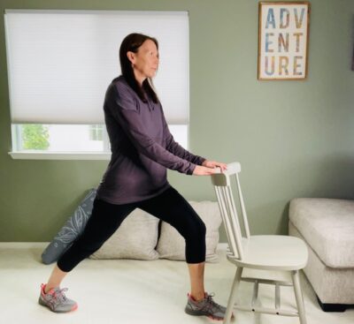 A woman practicing standing yoga positions with a chair for support with one foot forward and bent for a high lunge.