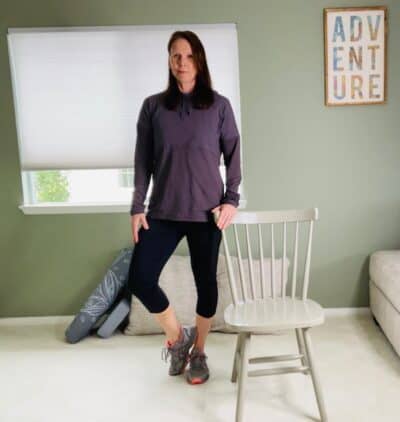 A woman practicing standing chair yoga positions supported by a chair with one foot planted and the other lifted up on the toe for a tree pose variation.