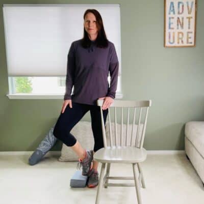 A woman practicing tree pose with the support of a chair and resting the foot on a yoga block.