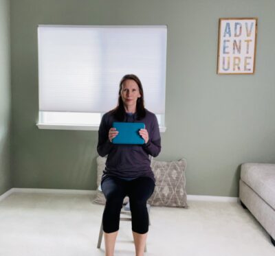 A woman seated in a chair holding a blue yoga block with both hands near her chest.