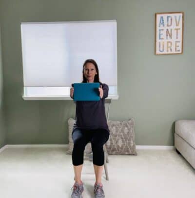 A woman seated in a chair holding a yoga block with both hands stretched out in front of her body.