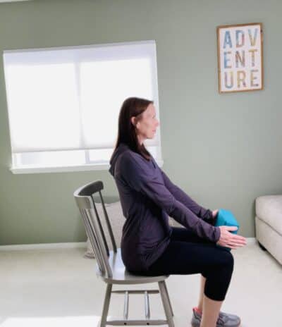 A woman seated in a chair and holding a blue yoga block resting on her knees between her hands.