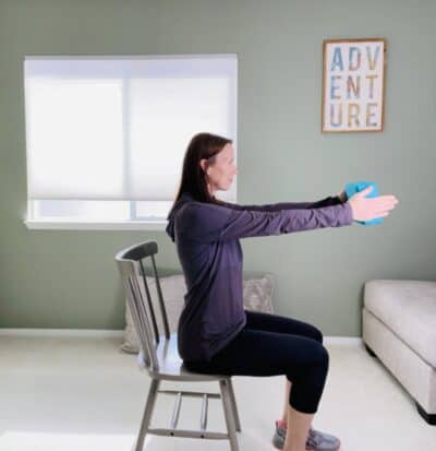 A woman seated in a chair and holding a blue yoga block between her hands as she raises the block up to shoulder height.