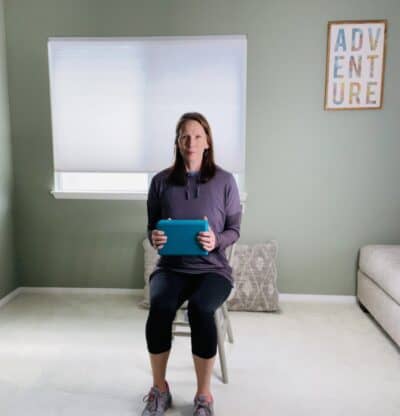 A woman seated in a chair holding a blue yoga block with both hands in front of her body.