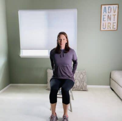 A woman seated in a chair and holding a yoga block with both hands behind her body.