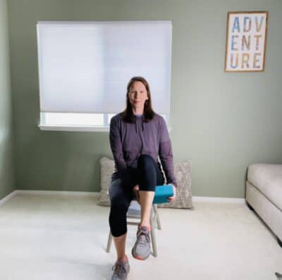  A woman seated in a chair passing a yoga block from one hand to another under the thigh of one lifted leg.