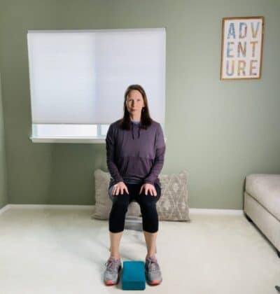 A woman seated in a chair with a blue yoga block on the floor between her feet.