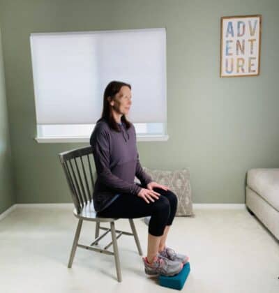 A woman seated in a chair with her feet resting on a yoga block and her heels reaching toward the floor,