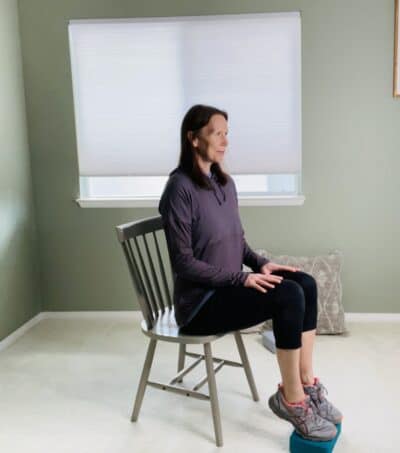 A woman practicing heel raises on a yoga block while seated in a chair.