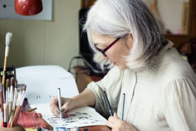 A cropped image of an older woman with a white sweater and glasses seated at a table and sketching with a pencil on a sketch pad.