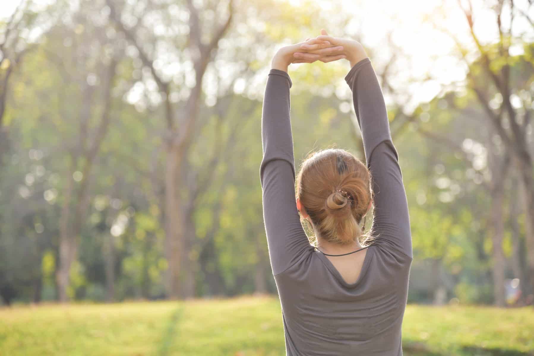 A woman with her hands clasped together and stretched over her head outside in a park for a shoulder exercise for flexibility.