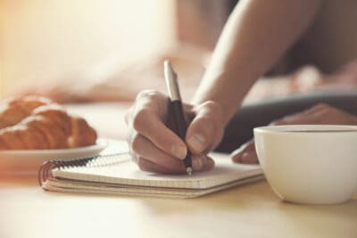 A cropped image of a woman writing in a journal with a pen on a table with a coffee cup.