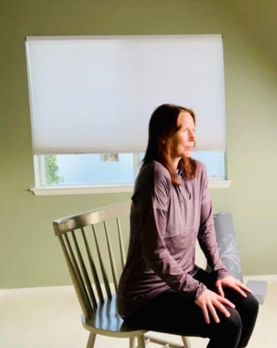 A woman seated in a chair with her shoulder rolled forward for chair yoga exercise shoulder circles.