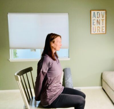 A woman seated in a chair with her hands clasped together behind her body for modified chair yoga pose, bound hands stretch.