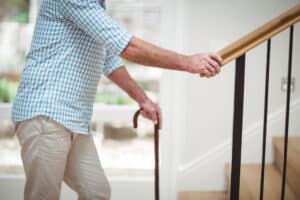 A cropped picture of an older man climbing stairs in a home with one hand on the railing and the other hand holding a cane.