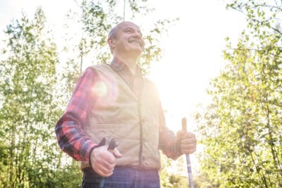 A cropped image of an older man holding walking poles and smiling as her walks in the sunlight outside in a tree filled area.