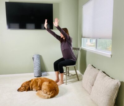 A woman seated in a chair near a window with her dog next to her practicing a modified downward facing dog with her arms raised in front of the body and her wrists extended.